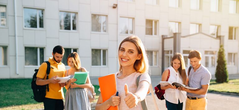 Smiling student giving a thumbs up outside a campus building representing success in higher education
