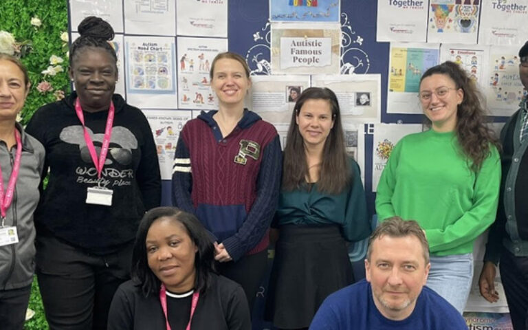 Group of Mont Rose College staff and students standing together in front of a decorated display board with posters and achievements, celebrating teamwork, diversity, and academic engagement, reflecting Mont Rose College’s 18 years Journey of Impact and Innovation