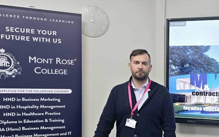 Martynas Masiulis posing beside a Mont Rose College banner and presentation screen during the Inspiring Guest Talk with Martynas Masiulis.