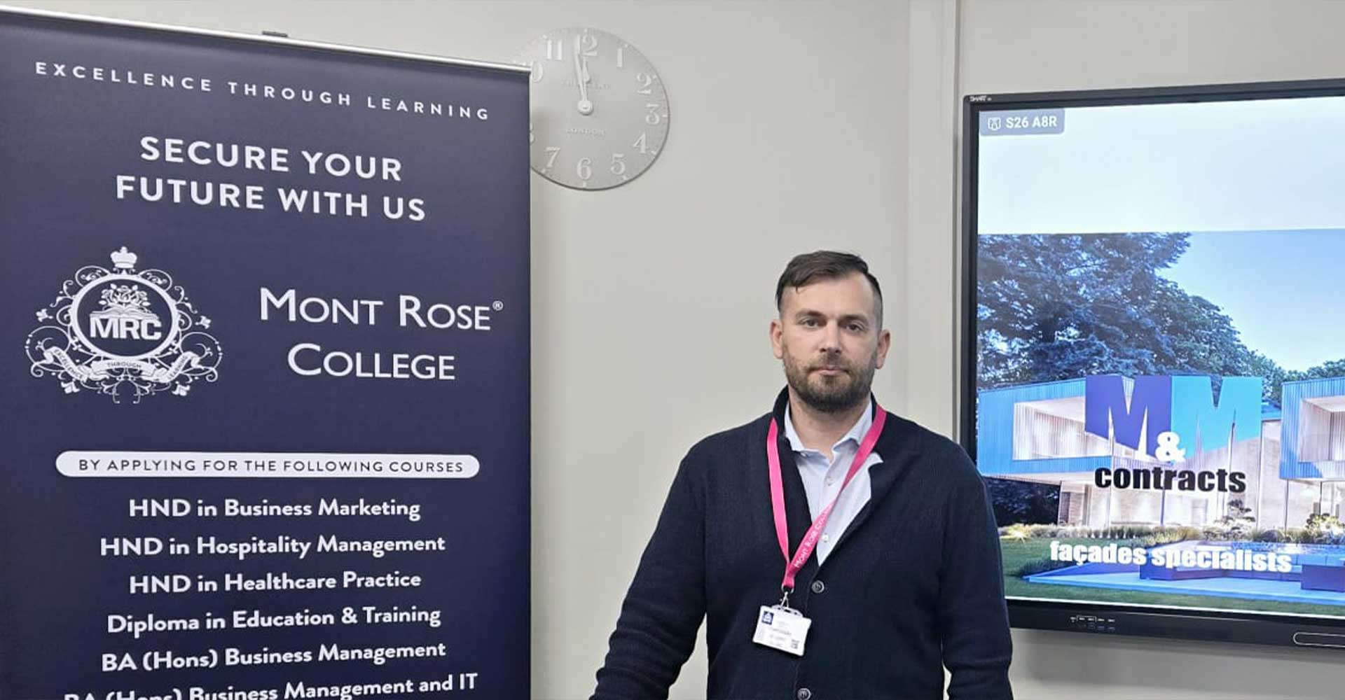 Martynas Masiulis posing beside a Mont Rose College banner and presentation screen during the Inspiring Guest Talk with Martynas Masiulis.