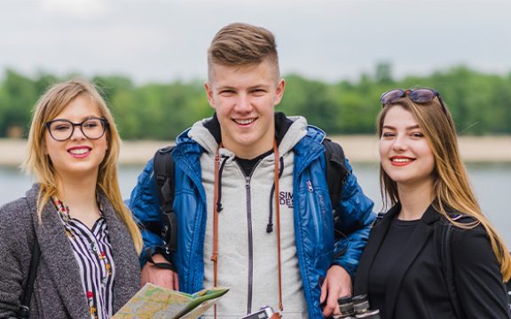 Three smiling young adults standing outdoors holding a map and camera by a body of water