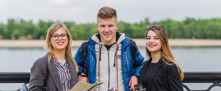 Three smiling young adults standing outdoors holding a map and camera by a body of water