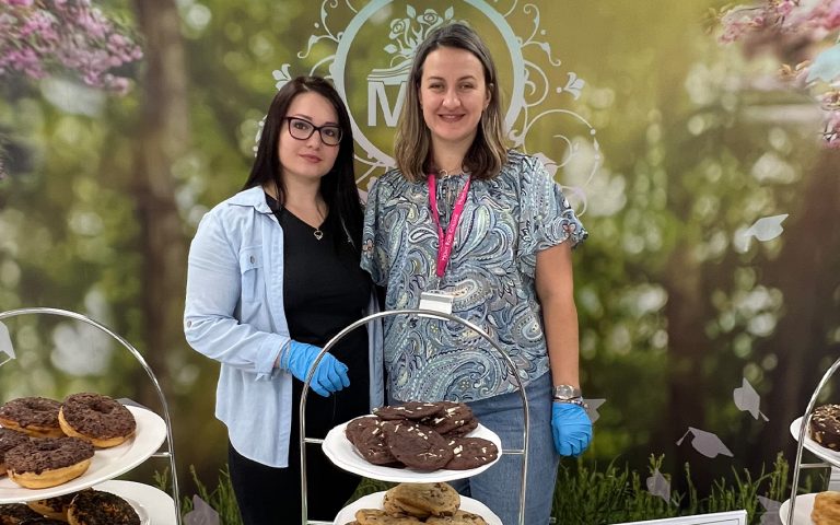 Two staff members posing at the Macmillan Coffee Morning at Mont Rose College, standing behind a dessert display with donuts and cookies, against a floral backdrop.