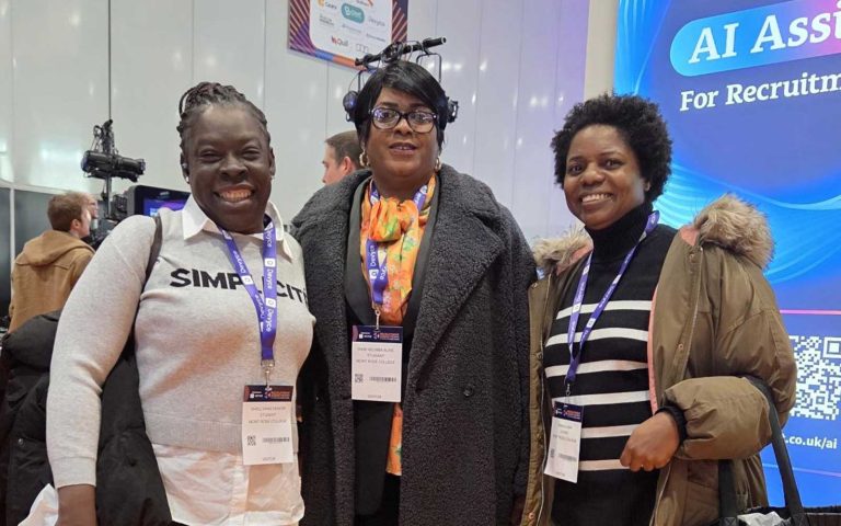 Three women smiling at the Recruitment Agency Expo, wearing visitor badges and standing in front of an AI Assist display.