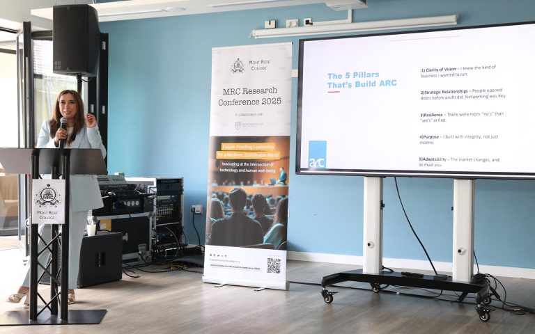 Speaker presenting at Mont Rose College podium during the MRC Research Conference, with a screen showing leadership pillars.