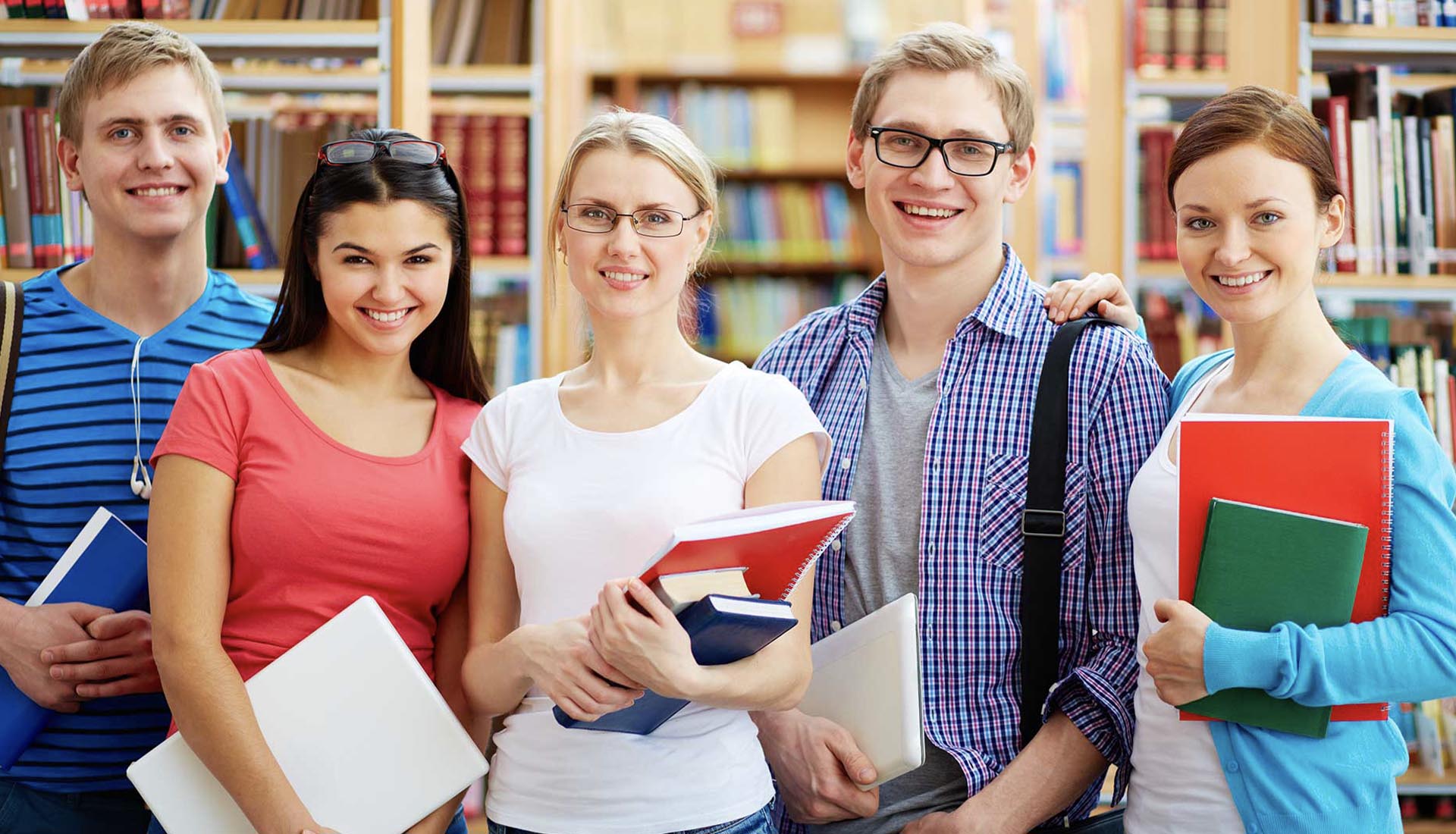 Group of students smiling in a library with books and laptops, symbolizing academic learning in HND vs On-the-Job Training.