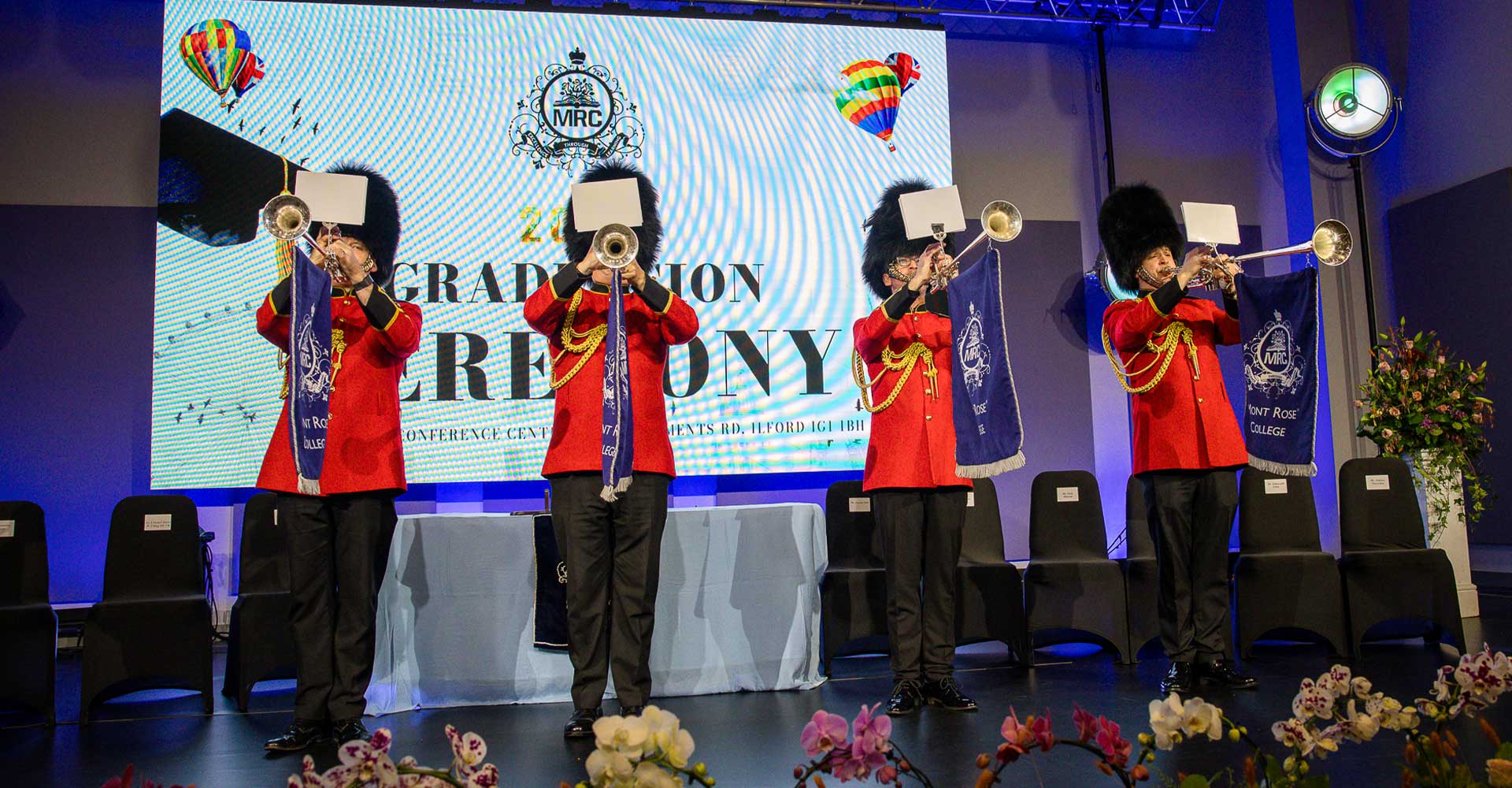 Uniformed trumpeters performing on stage during Mont Rose College Graduation 2025 ceremony, with college banners and floral decorations.