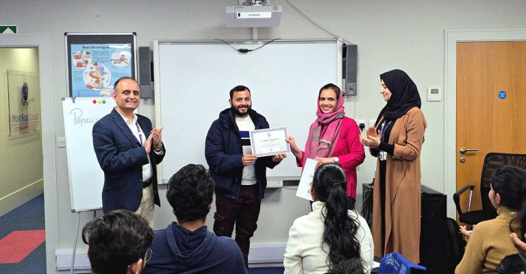 Participant receiving a certificate during the Strengthening Care Practice Through Physiological Measurements Workshop at Mont Rose College while attendees applaud in a classroom setting.