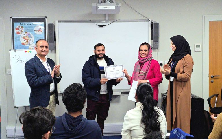 Participant receiving a certificate during the Strengthening Care Practice Through Physiological Measurements Workshop at Mont Rose College while attendees applaud in a classroom setting.