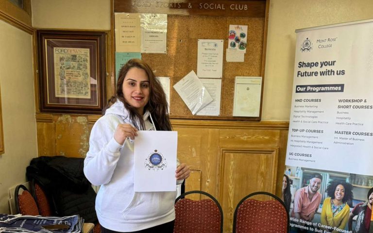 Woman representing Mont Rose College at the Lohri Community Event in Newbury Park, holding college materials beside a programme banner.
